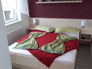 Uma cama com um cobertor vermelho e verde, duas almofadas, uma mesa de cabeceira com abajur e uma prateleira acima da cama.