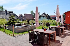 Un espace extérieur aménagé en terrasse, avec des tables et des chaises, un petit étang et, au fond, des bâtiments aux toits de chaume.