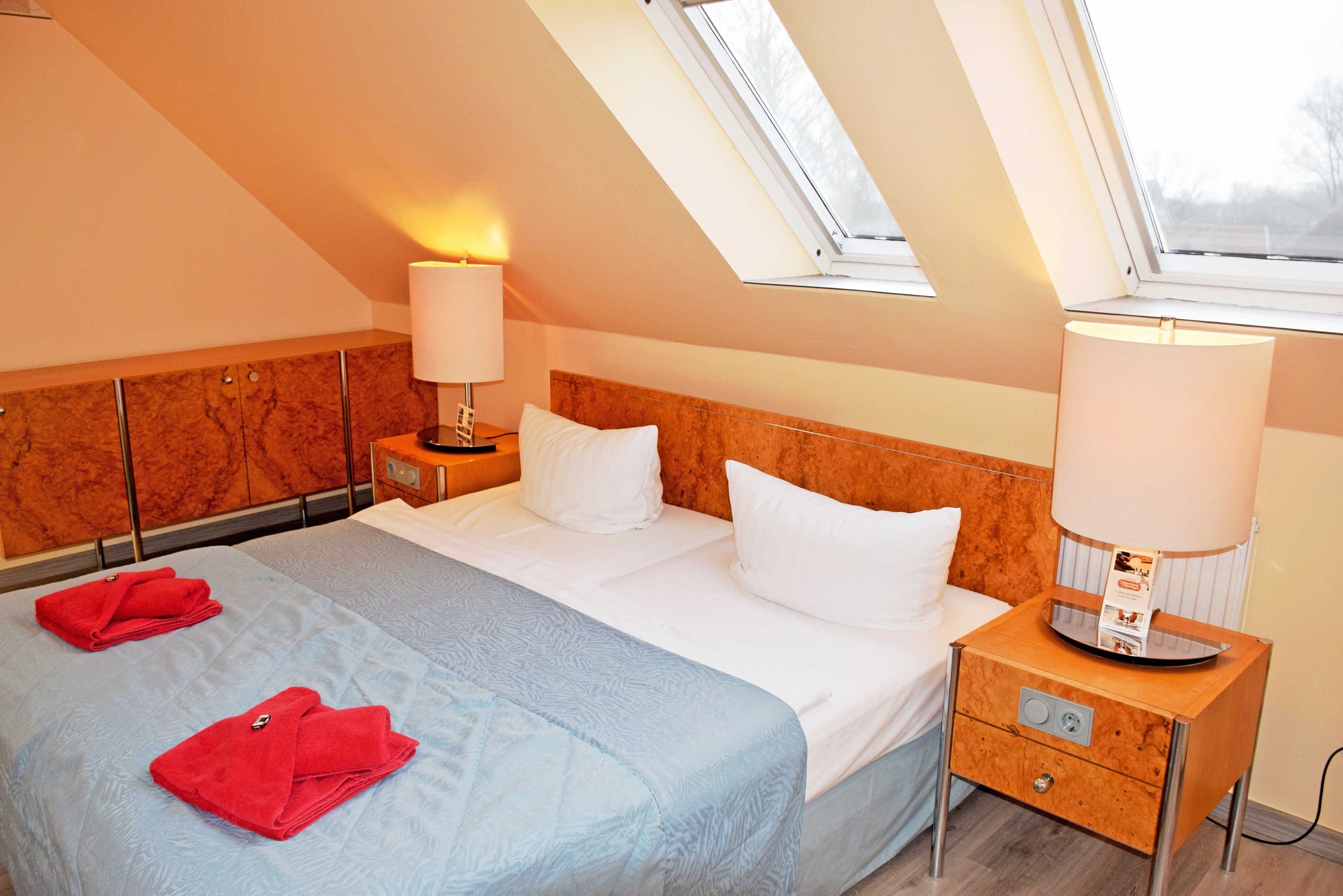 Une chambre d’hôtel avec un lit, deux tables de chevet, une petite table, une lucarne et une fenêtre.