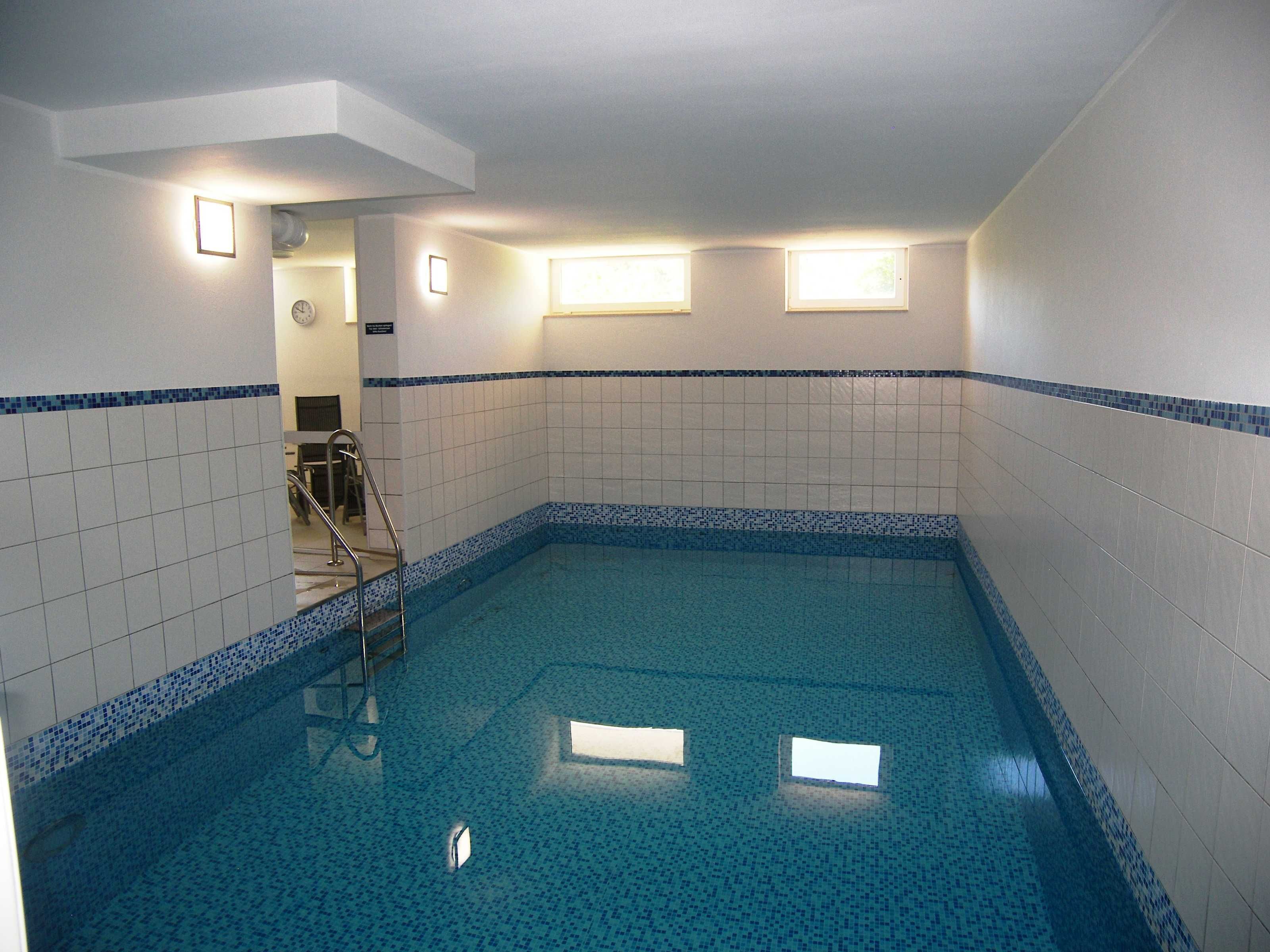 A small indoor pool with tiled walls and a blue border, a ladder, and a clock on the wall.