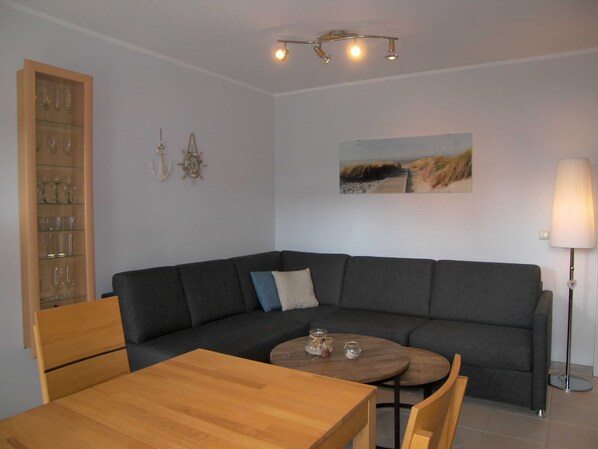A modern living room with a grey sectional sofa, a wooden dining table, and a round coffee table. There is a glass cabinet with wine glasses on the left and a lamp on the right.