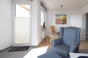 Une pièce avec une grande fenêtre, un fauteuil bleu, une chaise en osier et une table avec une lampe.