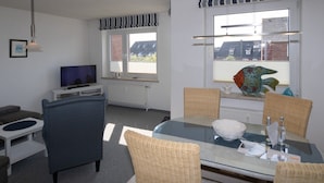 Ein modernes Wohnzimmer mit Fernseher, bequemen Sitzgelegenheiten, einem Esstisch und großen Fenstern mit Vorhängen.