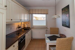 Une cuisine avec des meubles blancs, un pare-feu en carreaux, une fenêtre avec des stores, un luminaire suspendu et une chaise en osier.