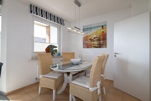 Un coin repas comprenant une table ronde en verre, quatre chaises en osier, une fenêtre agrémentée d’une lambrequin à rayures, un tableau représentant un paysage côtier et une porte.