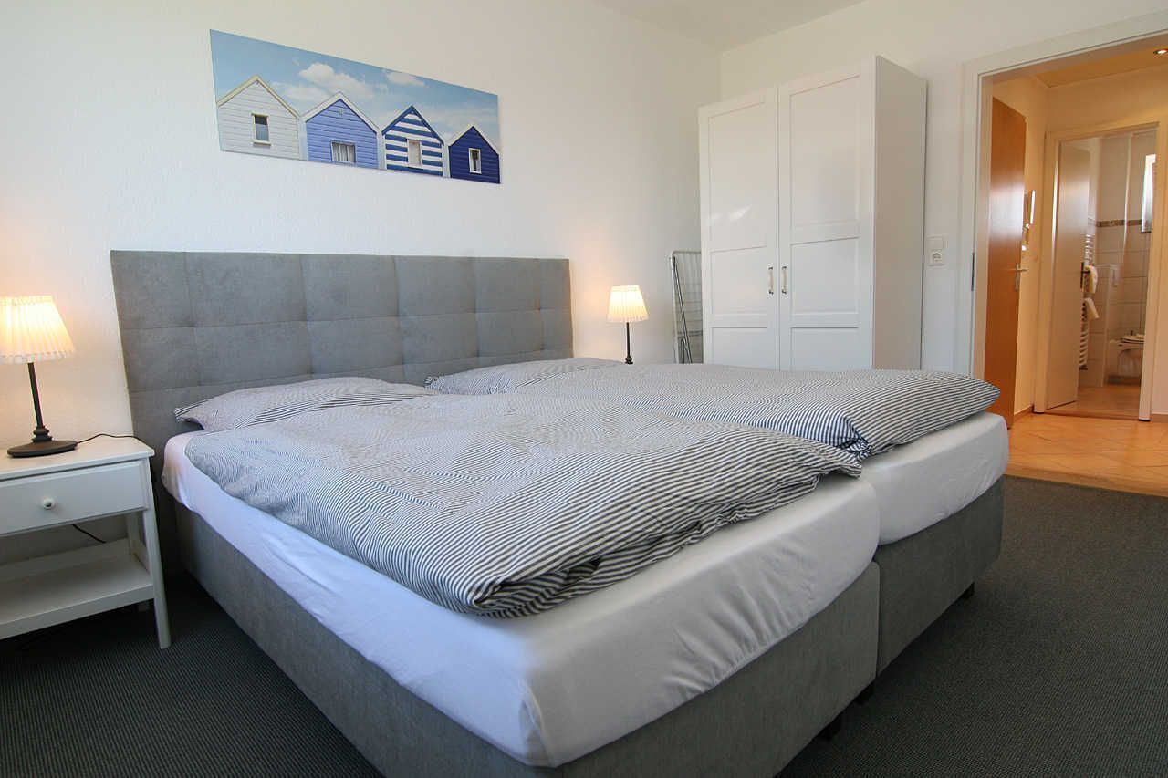A double bed with a grey headboard, two bedside tables with lamps, a painting of houses on the wall, and a door leading to a bathroom.