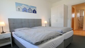 Ein Doppelbett mit grauem Kopfteil, zwei Nachttische mit Lampen, ein Hausgemälde an der Wand und eine Tür zu einem Badezimmer.