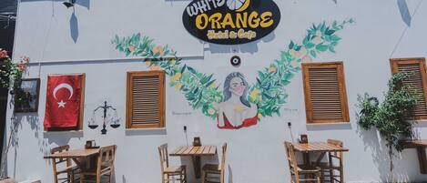 A building with a sign for "White Orange Hotel & Cafe" featuring a mural of a woman surrounded by leaves. There are two wooden tables and chairs outside.