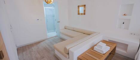 A modern, minimalist room with a beige sofa, a wooden table with towels, a glass door leading to a shower area, and a small mirror on the wall.