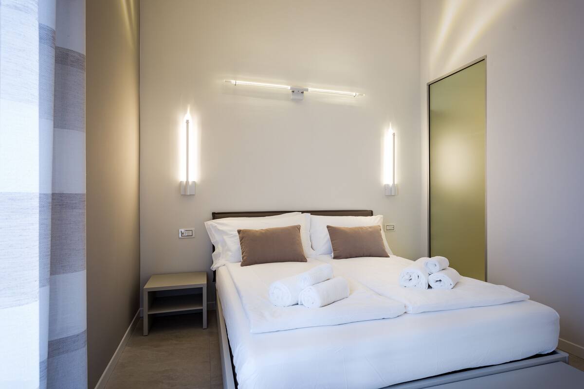 A neatly made bed with white linens and rolled towels, a bedside table, and wall-mounted lighting.