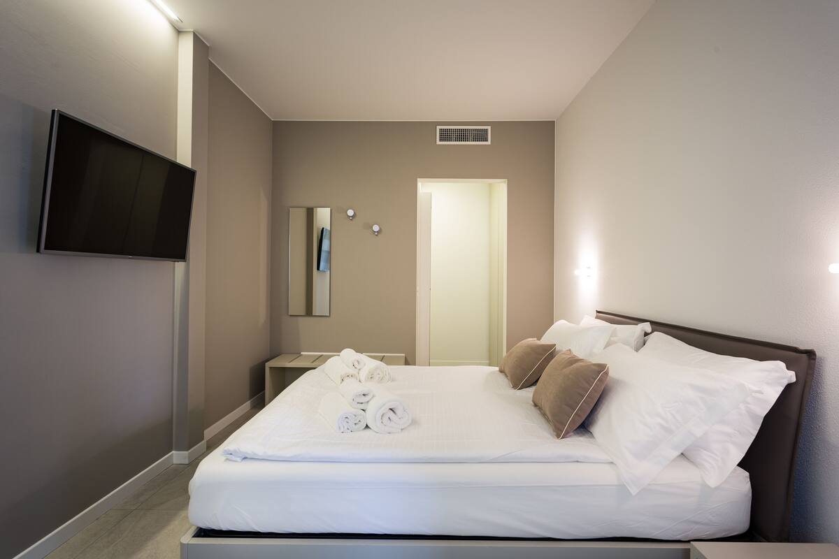 A modern hotel room with a large bed, a flat-screen TV mounted on the wall, a bedside table, and a door leading to another room.