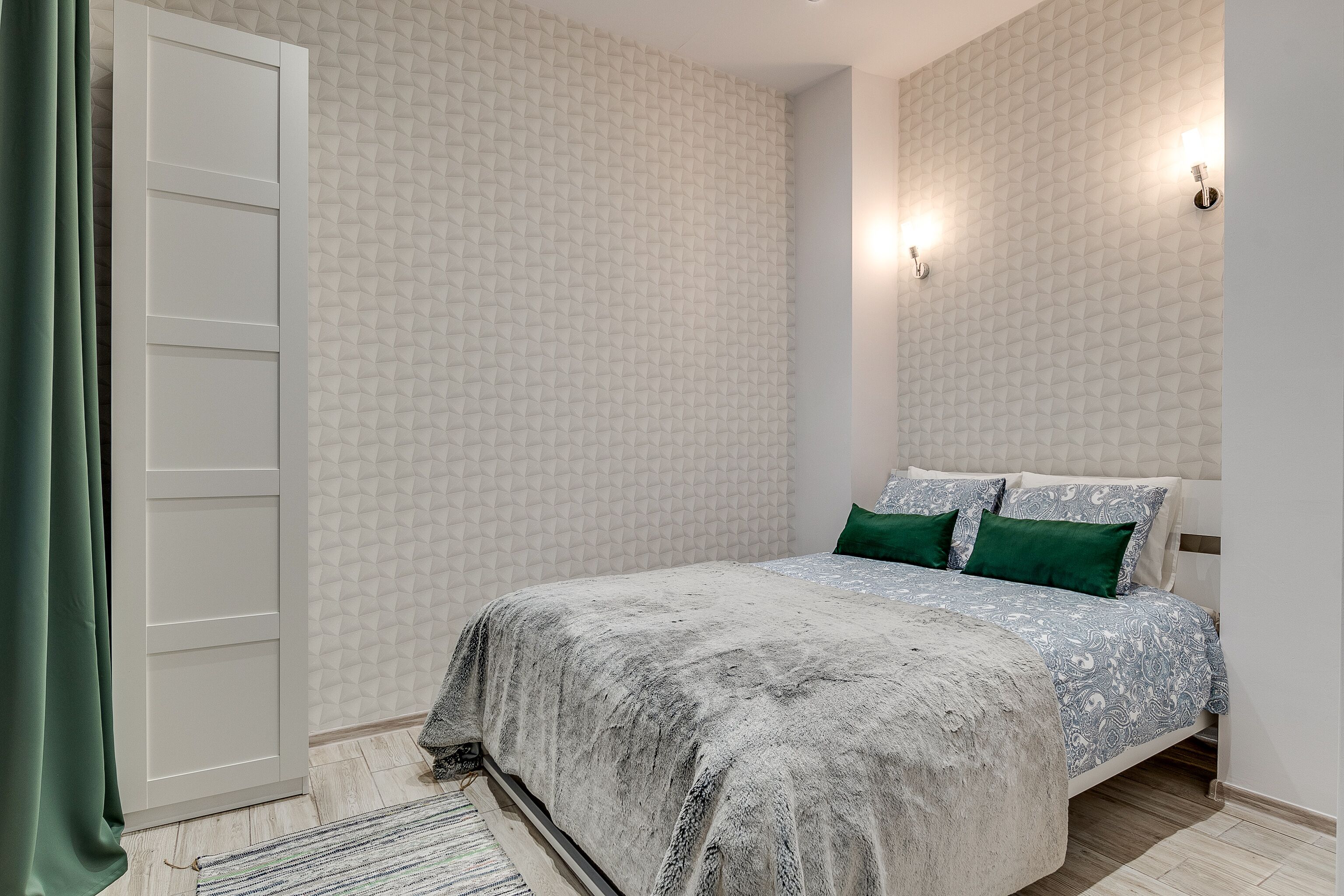A bedroom with a bed, green pillows, a patterned bedspread, a white wardrobe, and a geometric wall pattern.