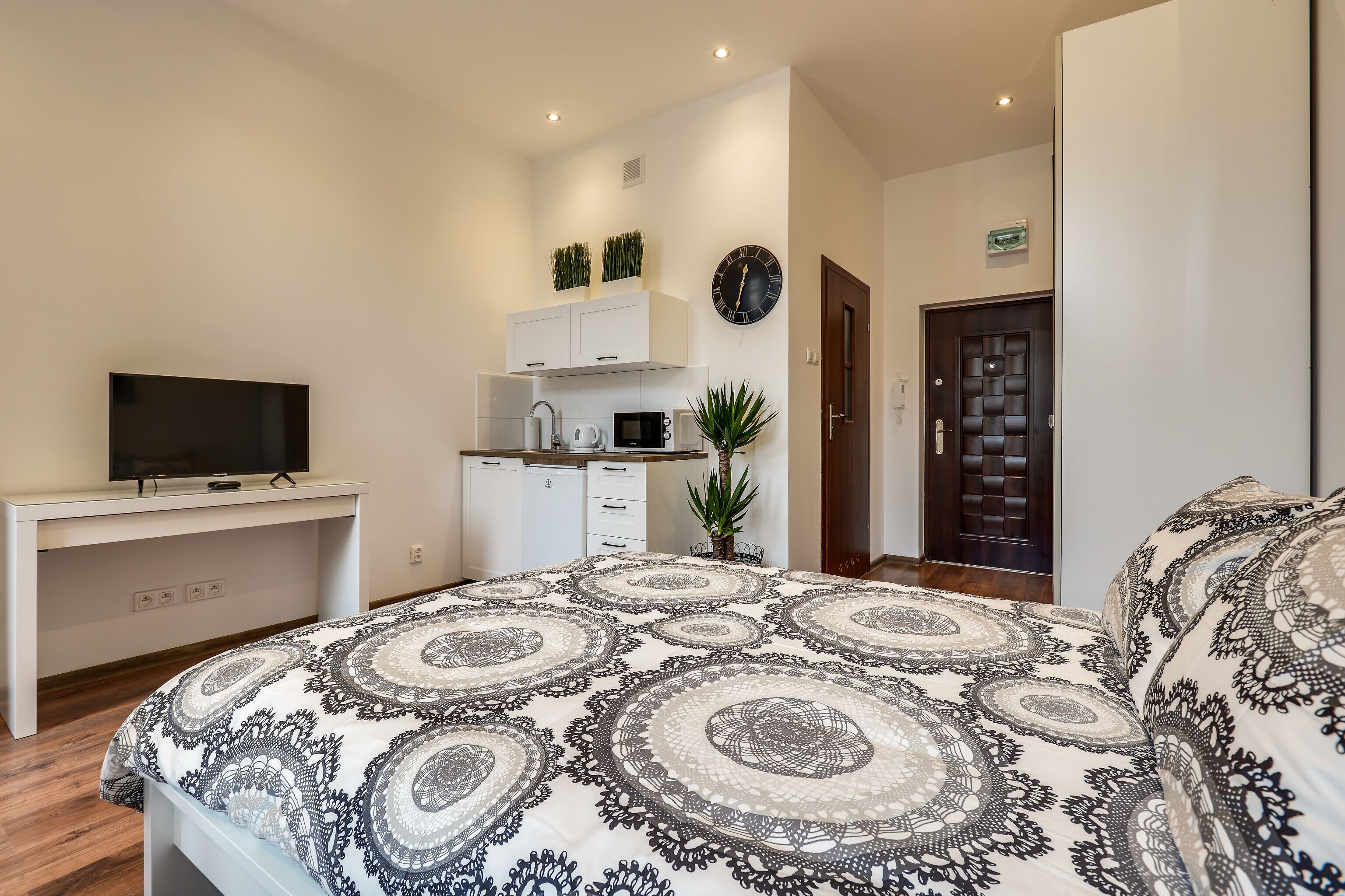 Ein Schlafzimmer mit einem gemusterten Bett, einem Fernseher auf einem Schreibtisch, einer Küchenzeile und einer Tür.