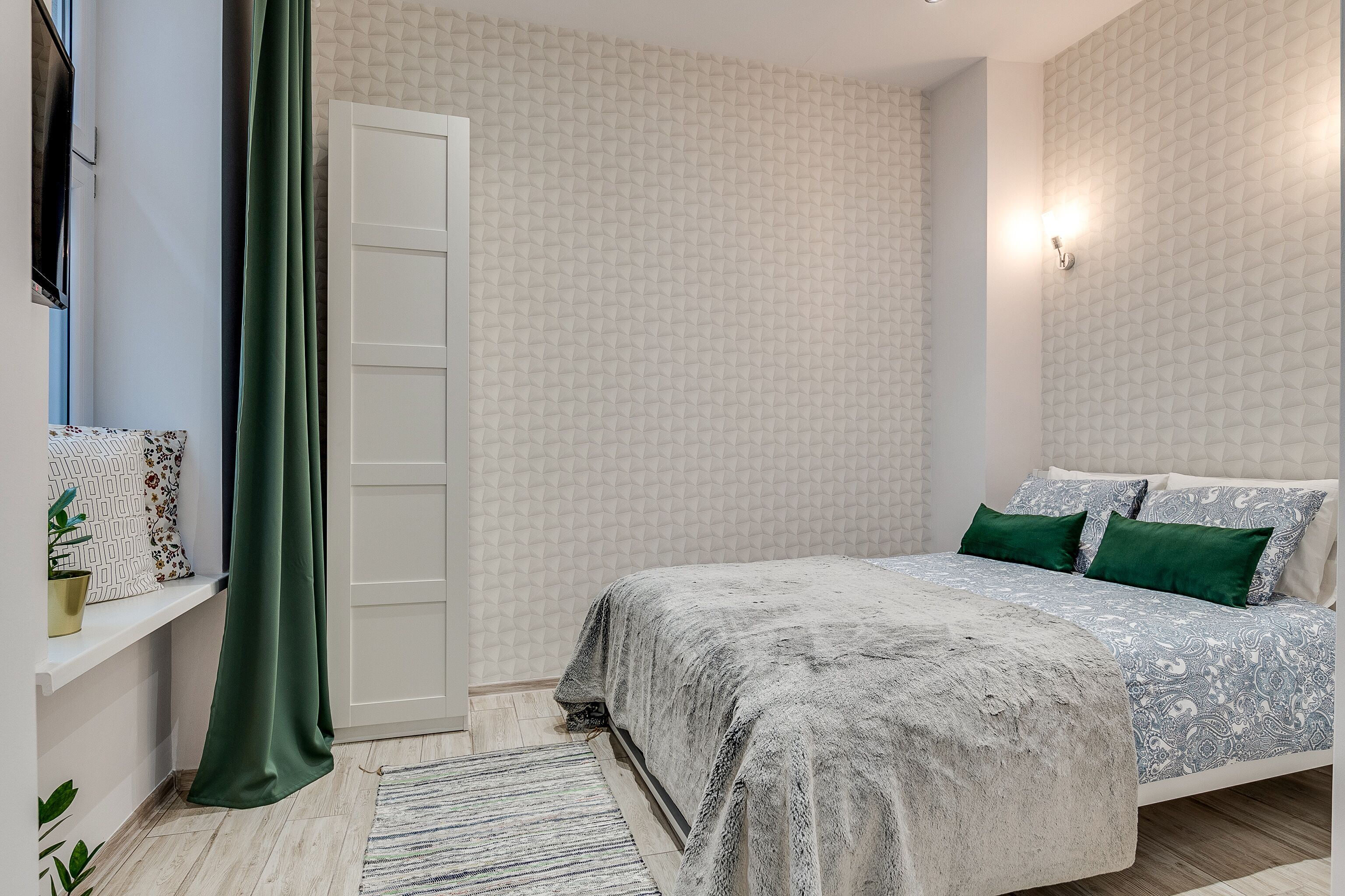 A bedroom with a bed, a window with a curtain, a wall-mounted TV, and a patterned wallpaper.