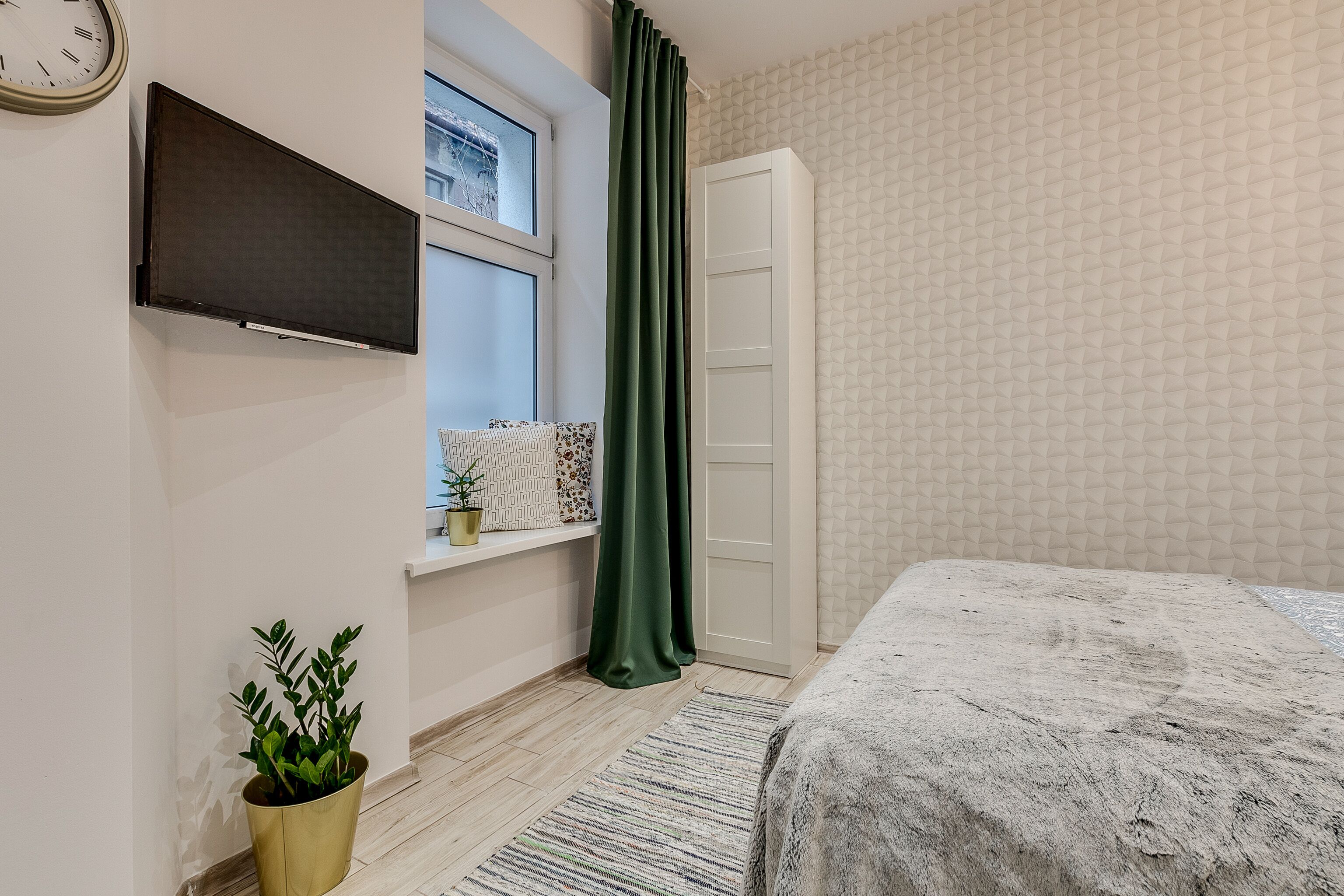 Quarto com cama, janela com cortinas, televisão na parede, planta em vaso e radiador.