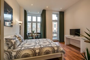 A bedroom with a bed, a desk, a TV, and a window with curtains.