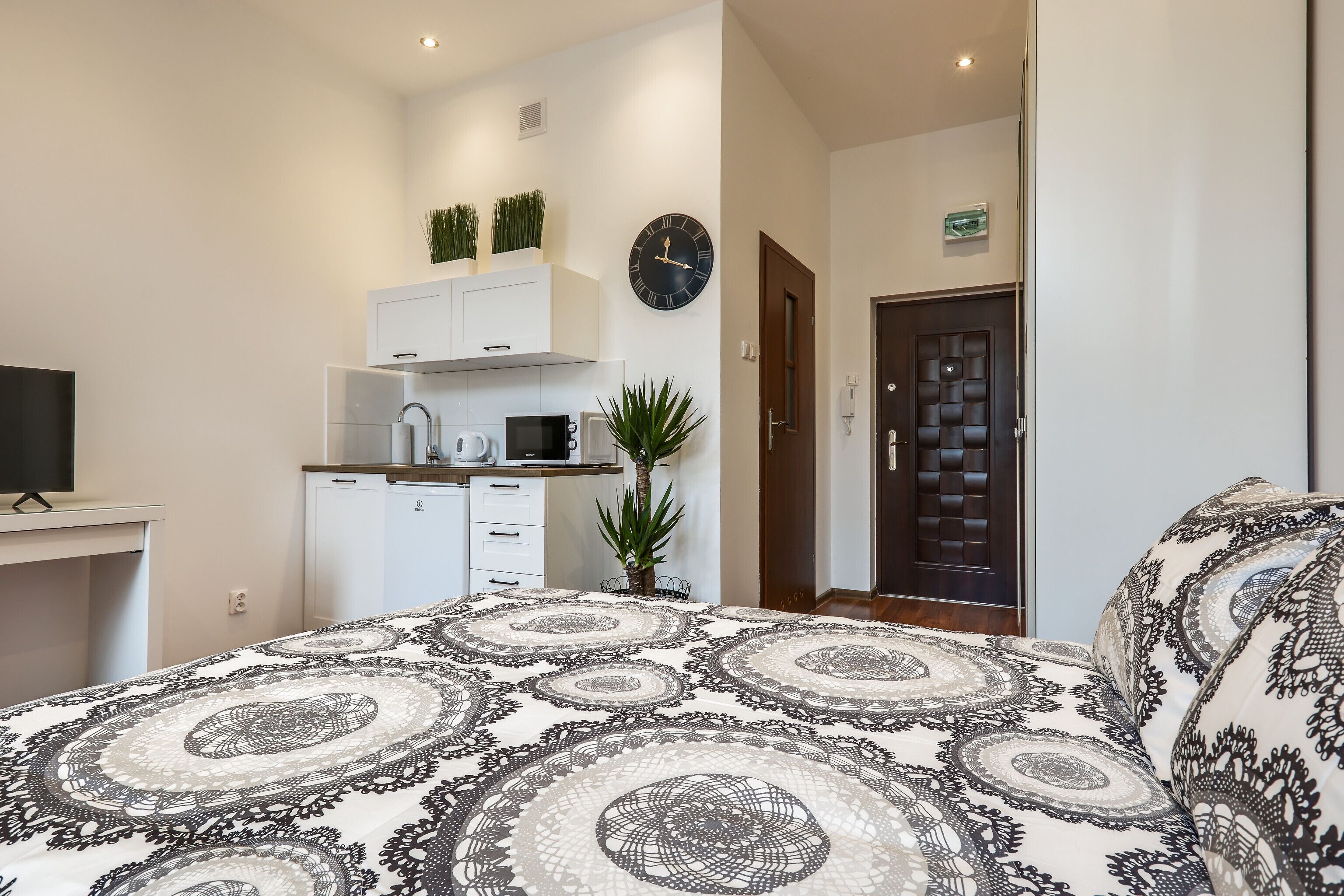 Quarto moderno com colcha estampada, pequena cozinha equipada com micro-ondas e um relógio na parede.