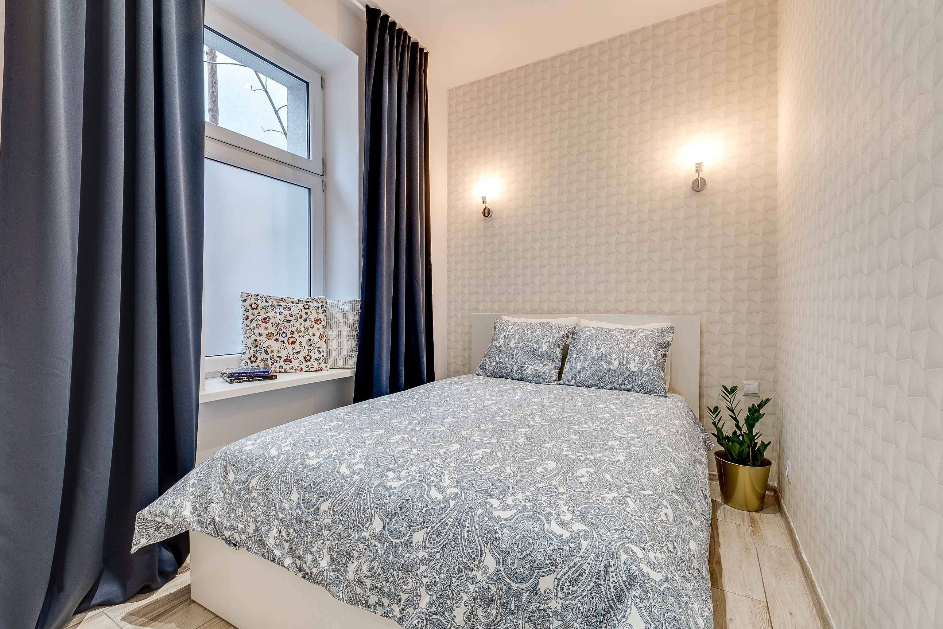 A bedroom with a large bed, a window with curtains, a bedside table with a book, and a wall-mounted light.