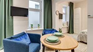 A compact living space with a sofa, a dining table with two plates, a TV mounted on the wall, and a bed with a headboard.
