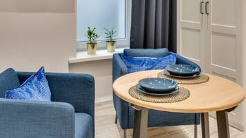 A modern living room with a round wooden table, two blue armchairs, and a sofa. There are two potted plants on a shelf and a window with curtains.