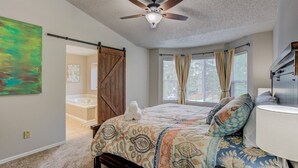 A bedroom with a bed, a ceiling fan, a large window with curtains, and a view of a bathroom. - 4BR AF Academyw/hot Tub & Firepit (Colorado Springs)