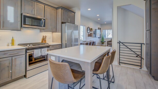 A modern kitchen with stainless steel appliances, a central island with bar stools, and a view to a dining area. - Modern Rooftop Patio New-build Townhome in COS (Colorado Springs)