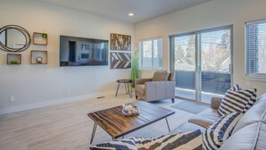 A modern living room with a flat-screen TV, a coffee table with decorative items, a sofa with striped cushions, and a large window with blinds. - Modern Rooftop Patio New-build Townhome in COS (Colorado Springs)