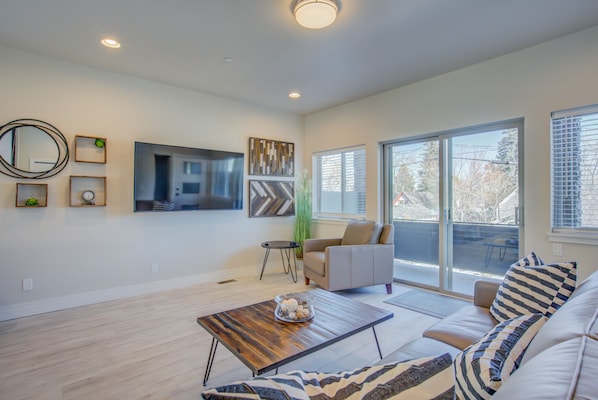 A modern living room with a flat-screen TV, a coffee table with decorative items, a sofa with striped cushions, and a large window with blinds.