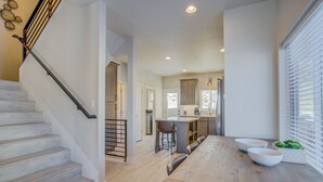 A modern kitchen with a central island, a dining area with a wooden table, and a staircase with a metal railing. - Modern Rooftop Patio New-build Townhome in COS (Colorado Springs)