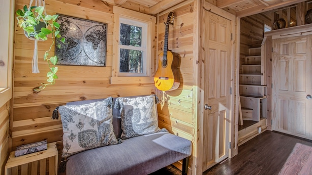 A cozy wooden cabin interior with a bed, a hanging plant, a guitar, and a staircase leading to another room.