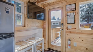 A wooden cabin interior with a kitchenette, a seating area with woven cushions, a door leading outside, and a mounted fire extinguisher.