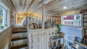 A wooden cabin interior with a bed, a kitchen area with pots and a stove, and a window with a view.