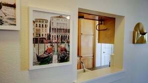 Une photographie encadrée représentant une scène de canal avec des bateaux et des bâtiments, un miroir reflétant une porte et un porte-manteau, ainsi qu’un petit coin lavabo.