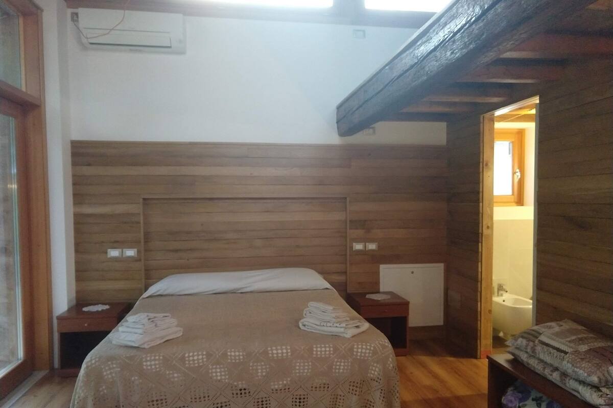 Un dormitorio con cabecera de madera, una cama con cubierta de encaje, una mesita de noche y un estante de madera con toallas dobladas.