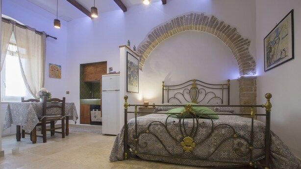 Un dormitorio con un arco de piedra, una cama con estructura metálica, una mesa de comedor con sillas y una zona de cocina.
