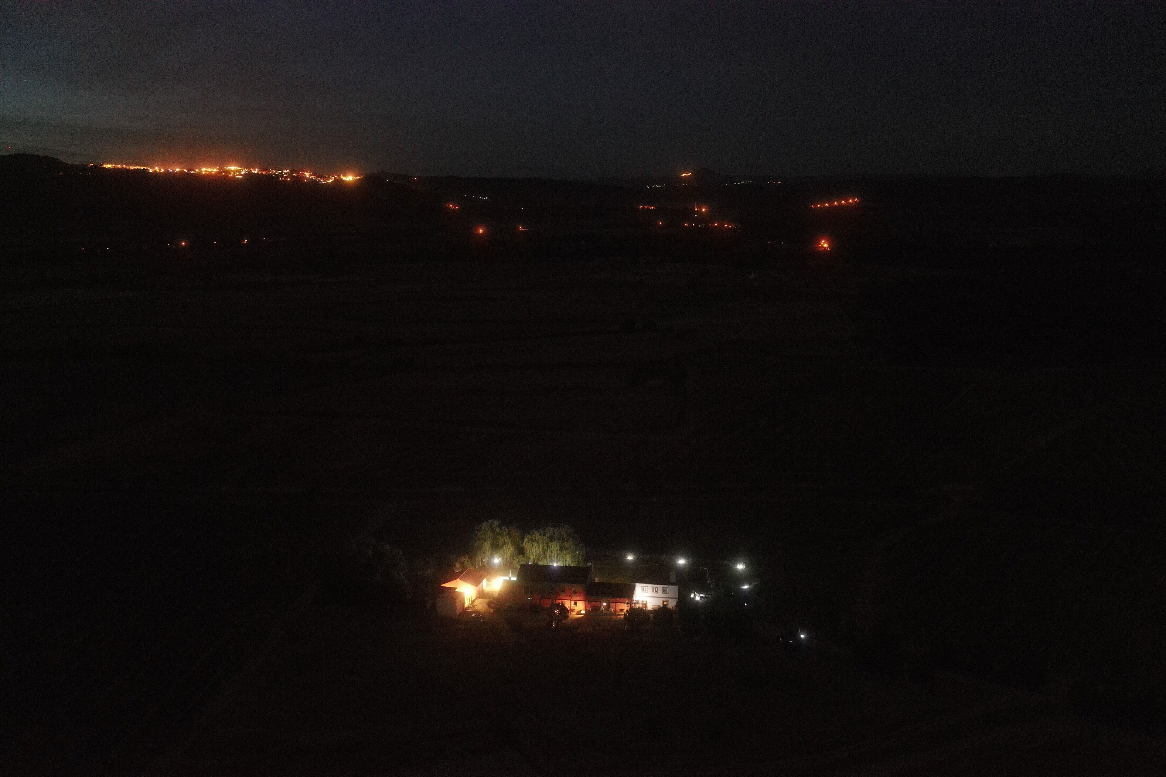 Vista nocturna de una zona rural con un pequeño conjunto de edificios, algunos con luces encendidas, y una hilera distante de luces al fondo.
