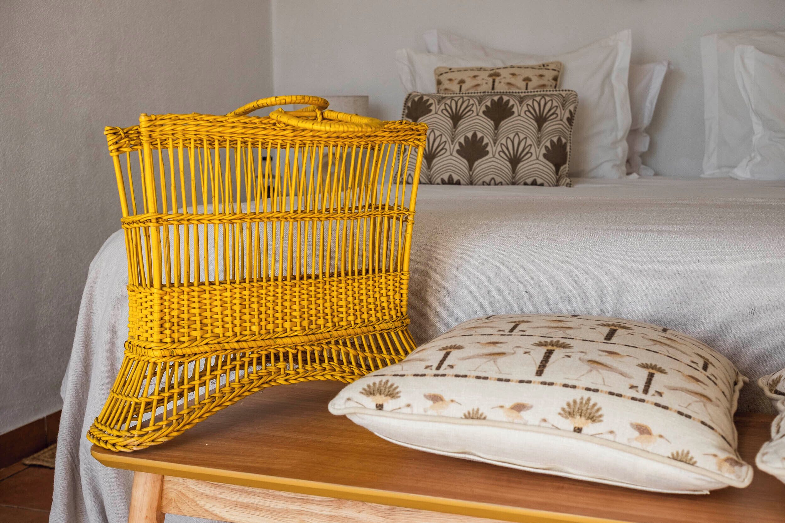 Una silla de mimbre con asa, un cojín estampado y una cama con ropa blanca.