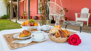 A breakfast setup with croissants, bread, fruit, and a red rose on a table outside a building with a pink wall and white chairs.