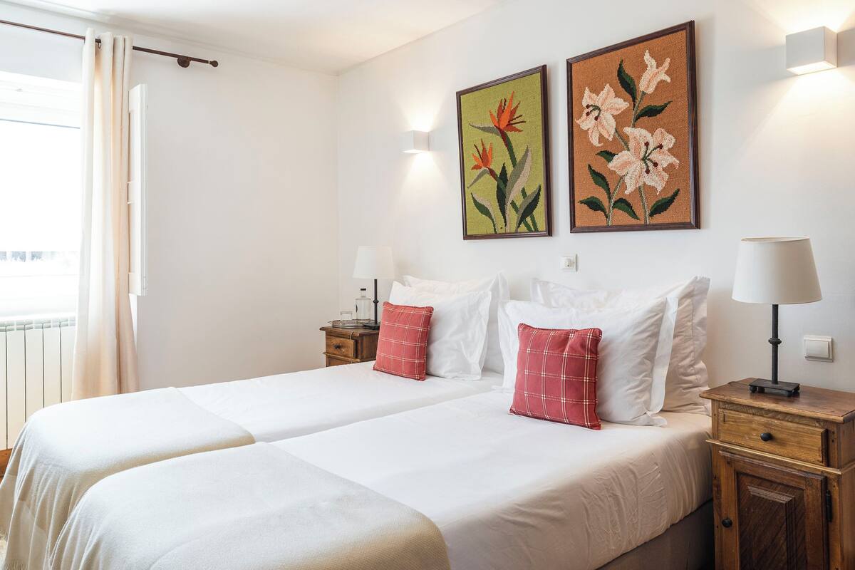 Un lit double avec une literie blanche et rouge, deux lampes fixées au mur et deux tableaux floraux encadrés.