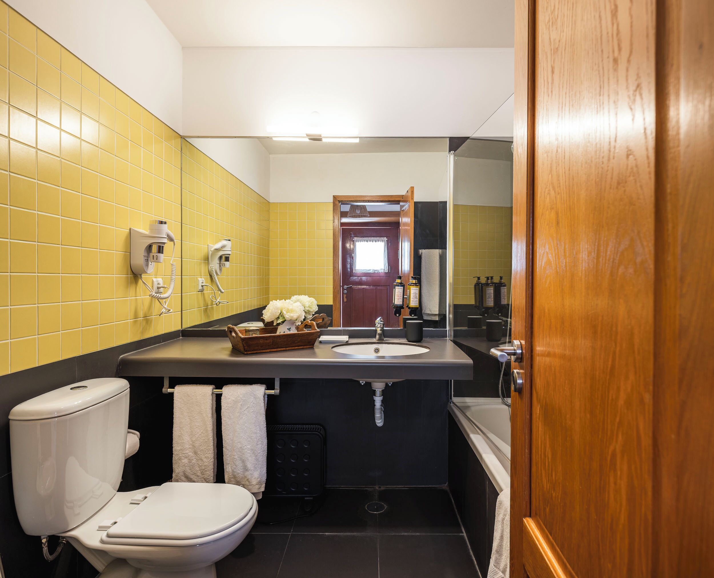 Un bagno con una parete rivestita di piastrelle gialle, un water bianco, un lavabo con uno specchio e una porta in legno.