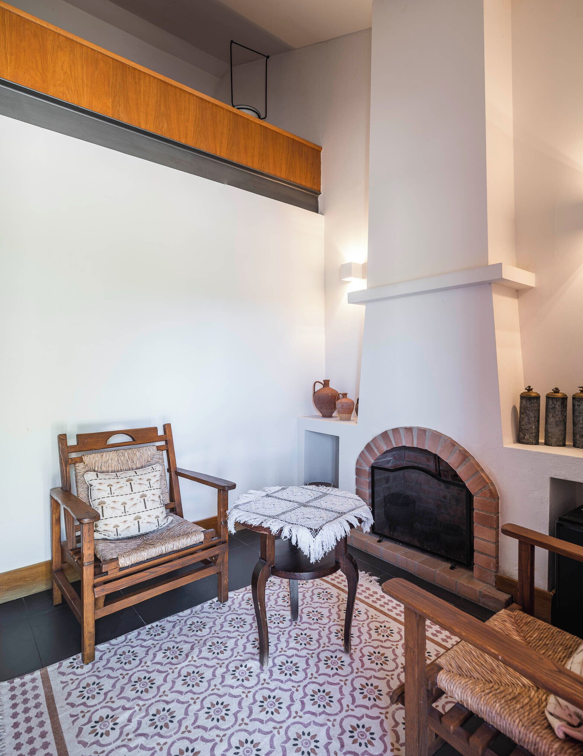 A cozy living room with a wooden chair, a small round table, a patterned rug, a fireplace, and a shelf with decorative items.