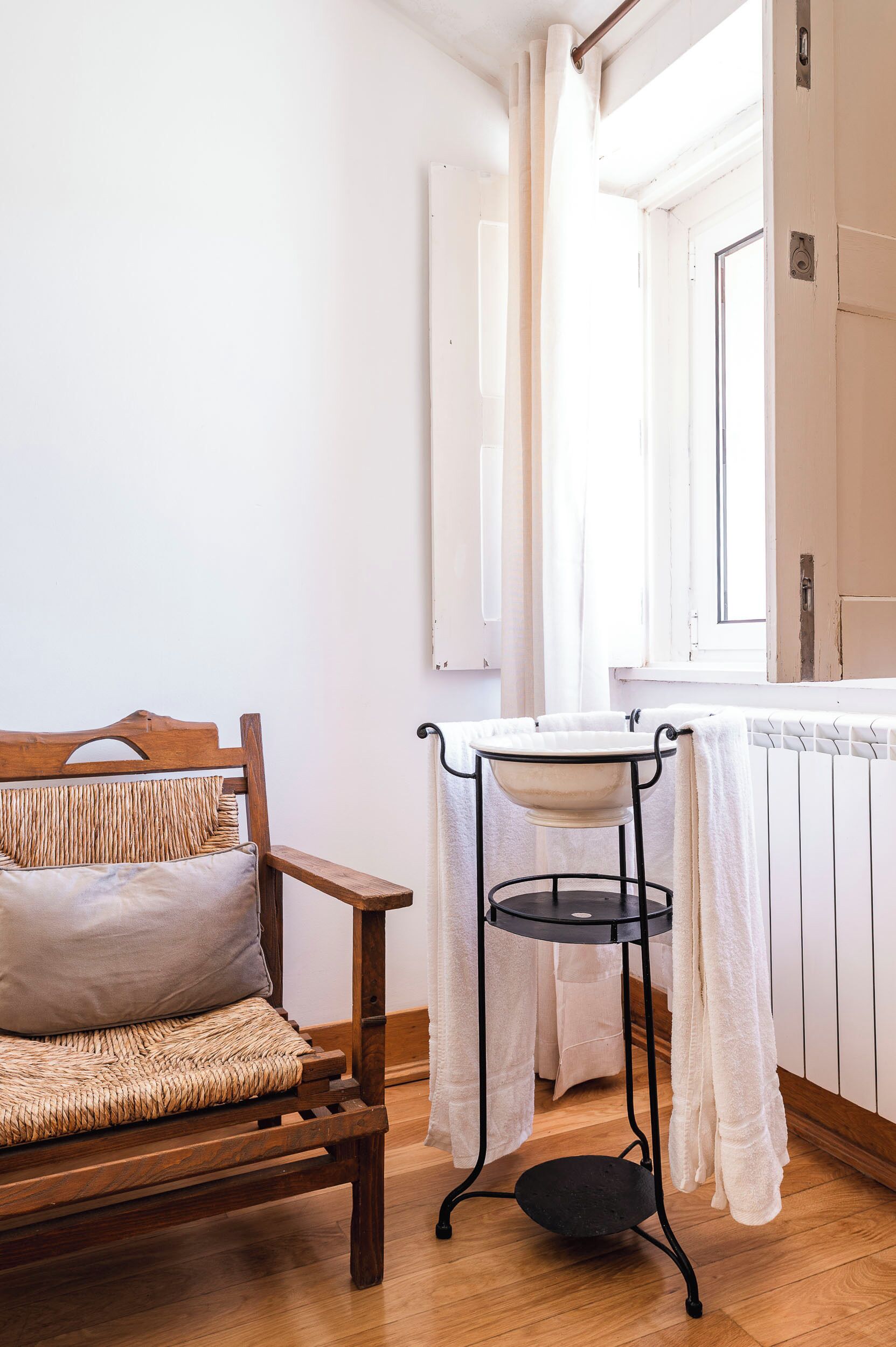 Un canapé en bois avec un coussin d’assise tissé, une table d’appoint en métal noir recouverte d’un tissu blanc et une fenêtre ornée de rideaux blancs.
