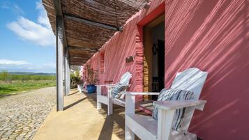 Un área de estar techada al aire libre con dos sillas blancas y un banco, un edificio rojo con una puerta, y un camino adoquinado.