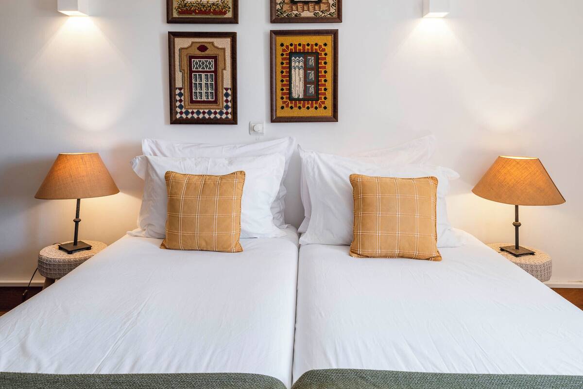 Un lit double avec du linge de lit blanc et deux oreillers assortis, de part et d’autre duquel se trouvent deux tables de chevet avec des lampes, et un tableau encadré est accroché au mur au-dessus.