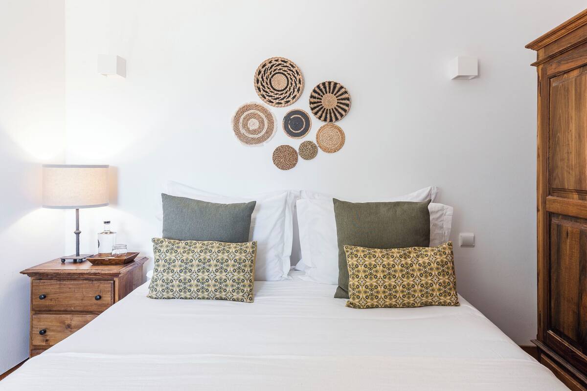 Un lit bien fait, agrémenté d’oreillers à motifs, une table de chevet avec une lampe et un objet décoratif fixé au mur.