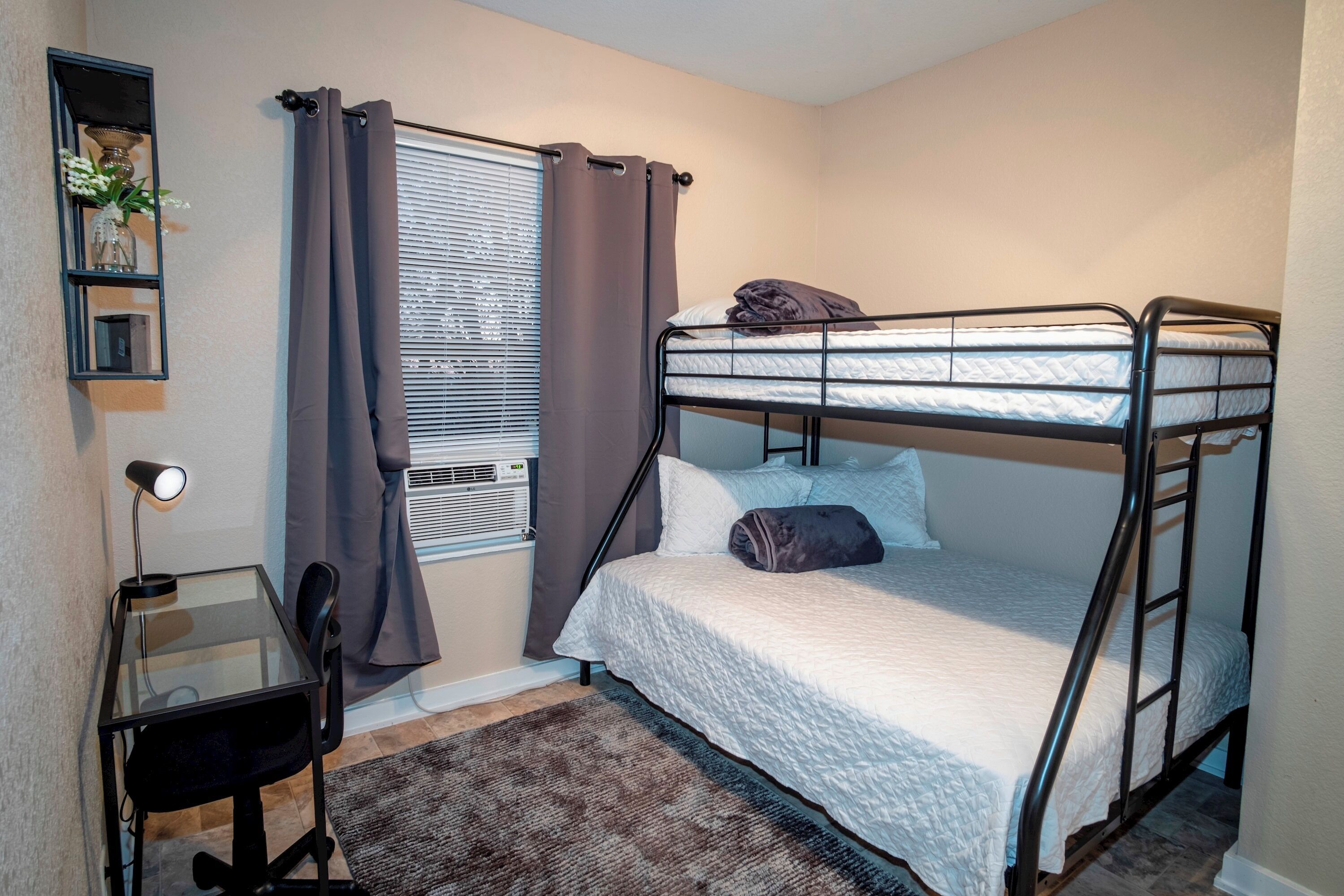 Une chambre compacte comprenant un lit superposé, un bureau avec une chaise, une petite étagère avec des objets décoratifs et une fenêtre avec des rideaux.