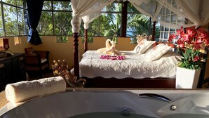 Um quarto luxuoso com uma cama grande, uma banheira com flores e vista para palmeiras através de janelas amplas.