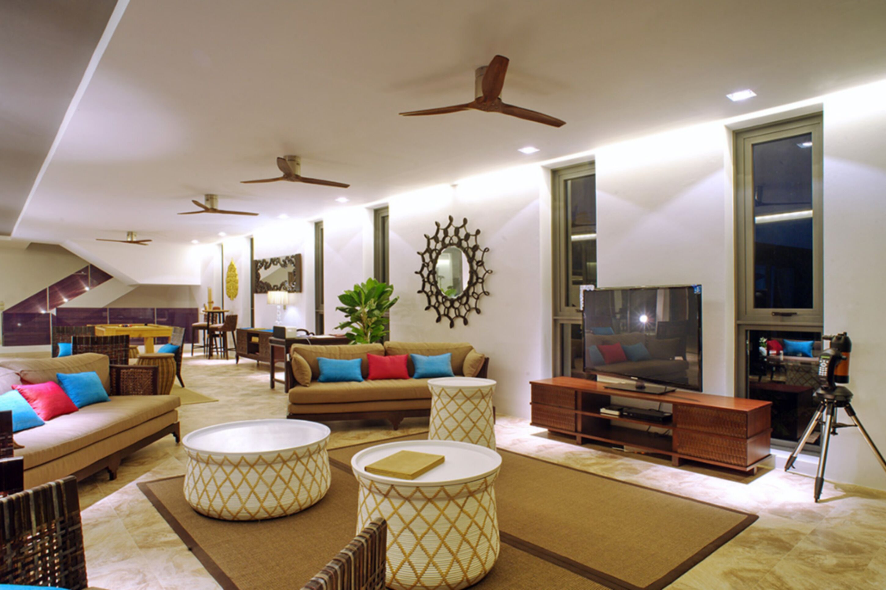 A spacious living room with a sofa, coffee table, and TV, featuring ceiling fans, large windows, and a plant.
