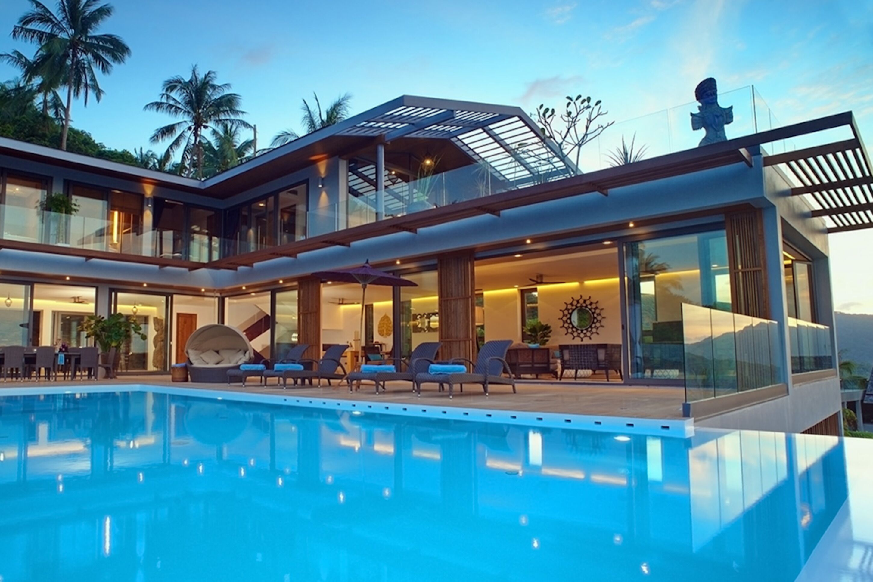 A modern multi-story hotel with a large pool, outdoor seating, and a tropical backdrop.