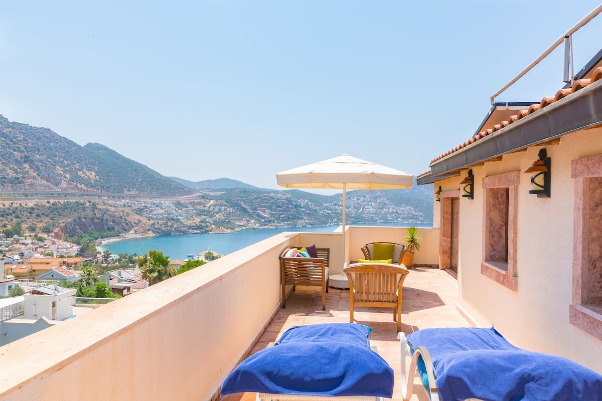 En balkon med to liggestole, en stol og en parasol med udsigt over en kystby og bjerge.